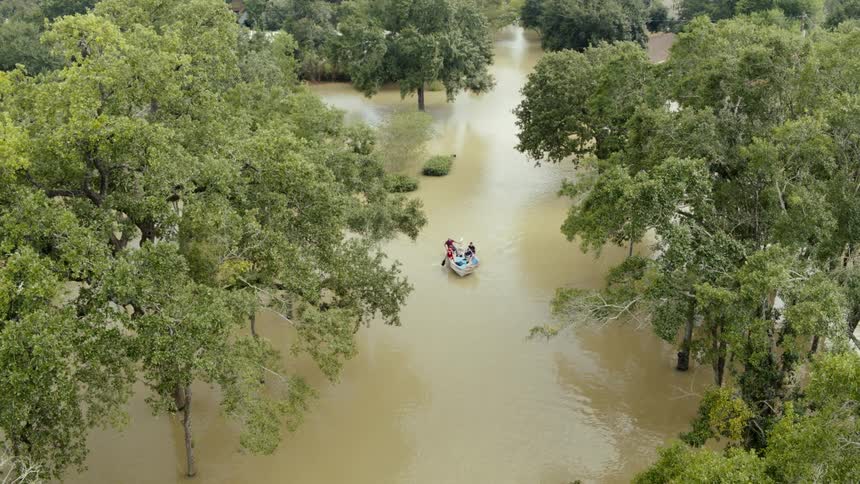 A boat is being paddled by two people as it moves down flood waters between trees with houses in the background