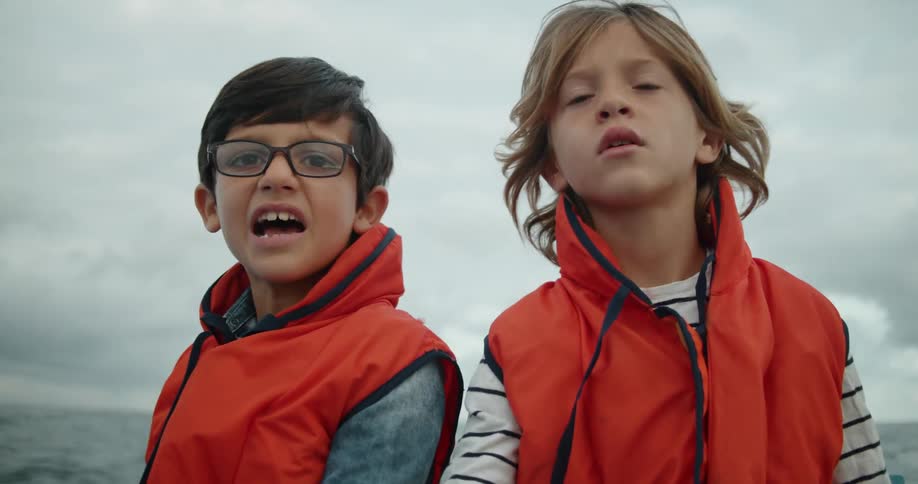 Kids wearing life vests sit in rocking boat under overcast sky