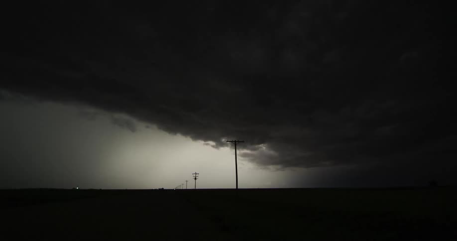 Lightning flashes in the clouds illuminating the dark night sky over a road lined with utility poles