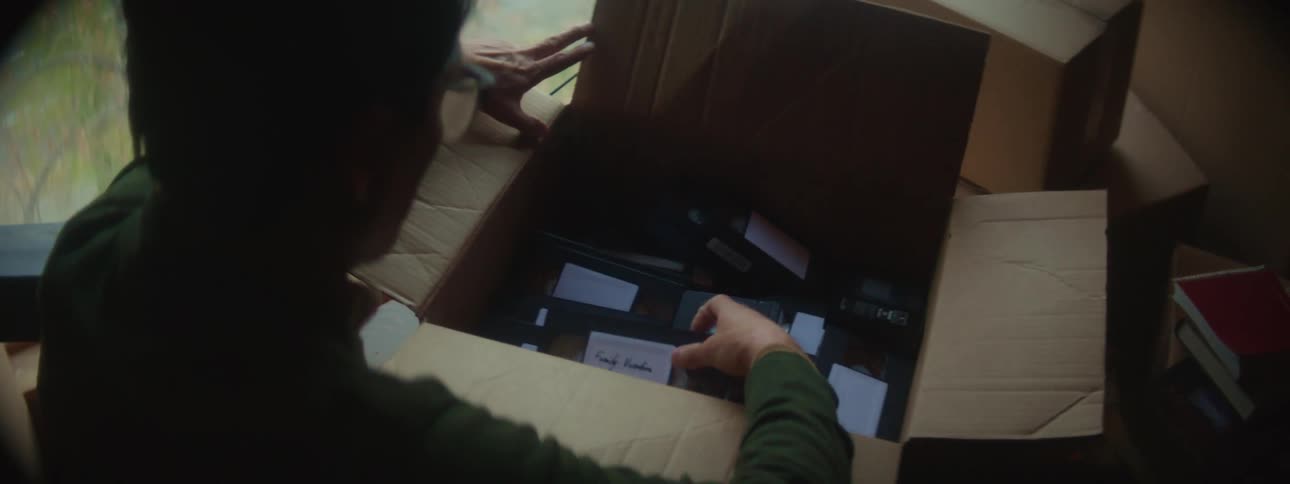 A person opens a cardboard box in a room and finds it full of vhs tapes