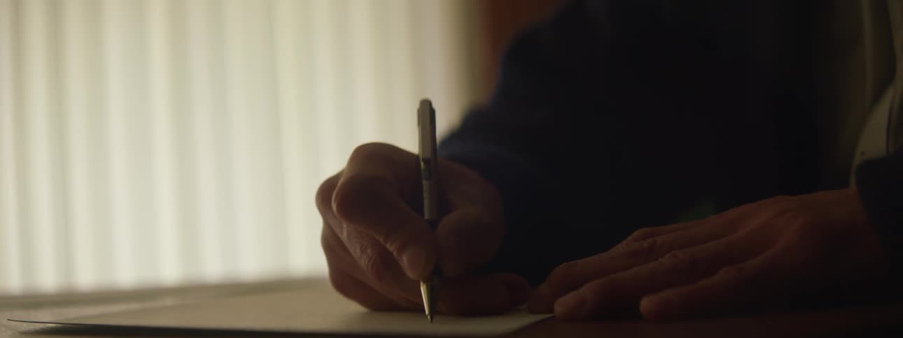 Man wearing jacket writes down in paper sitting at desk in room