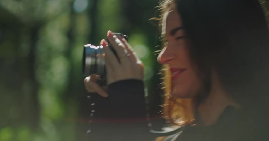 Woman standing among trees smiles while raising camera to her eye