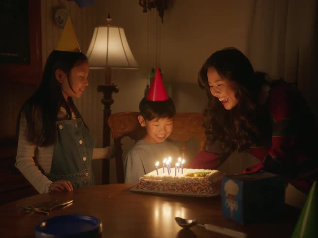 A mother carries a cake with candles to her son while the daughter is smiling and singing