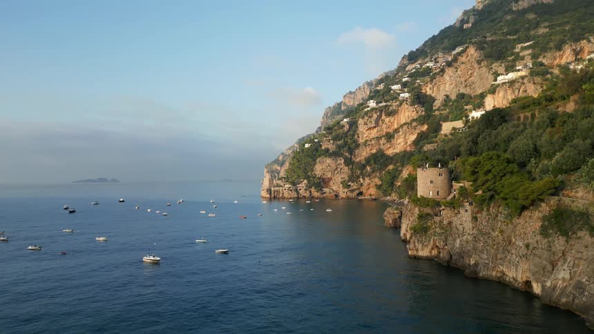 Many boats float off shore from seaside town and tower on mountain side
