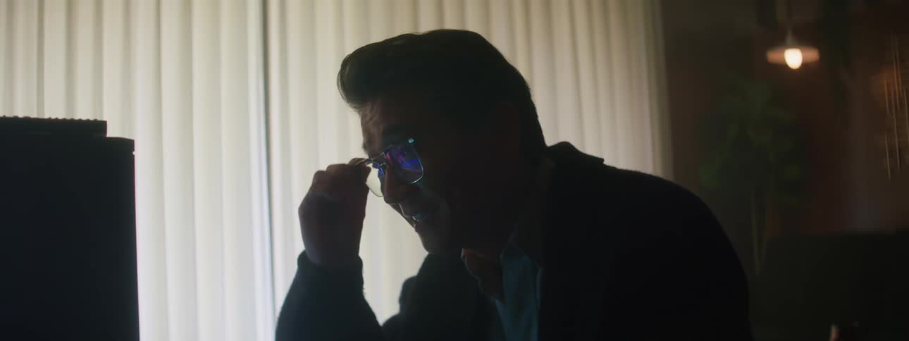 Close up of a man wearing glasses looking at a television screen on a dimly lit room at home