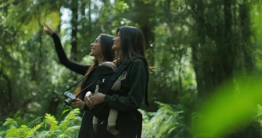 Two women are standing in the woods and one is holding her baby and the other is taking pictures