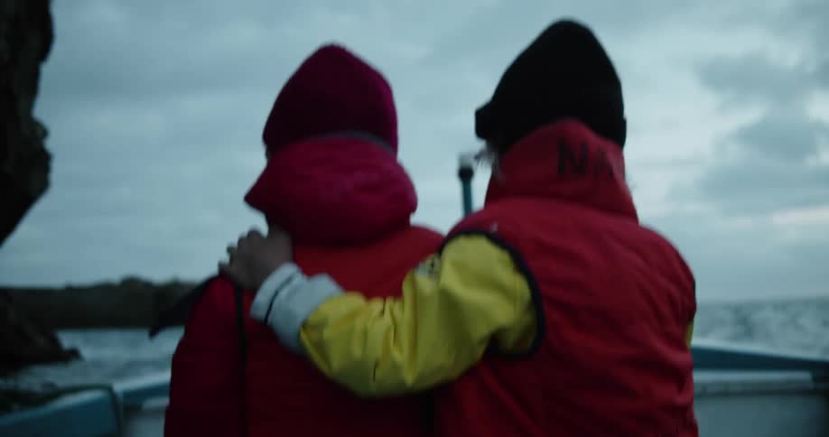 Two kids wearing life vests sit in bobbing boat near bottom of cliff