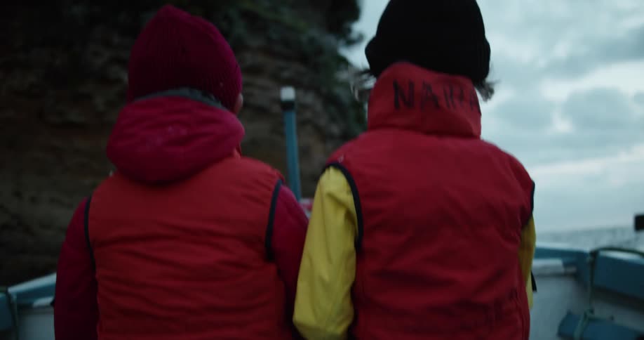 Two young boys wearing bright colorful jackets ride a boat close to the cliffs next to the ocean