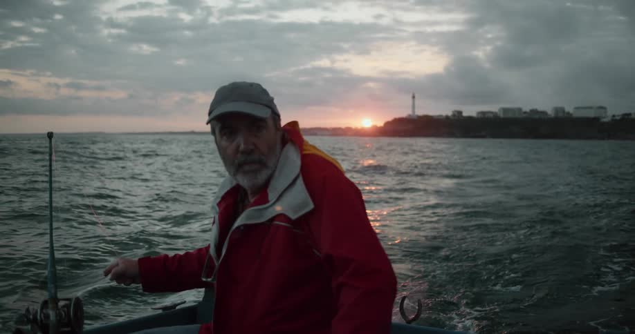 Man with brown cap and red jacket travels on boat floating on water with skyline of city and sunset in the background