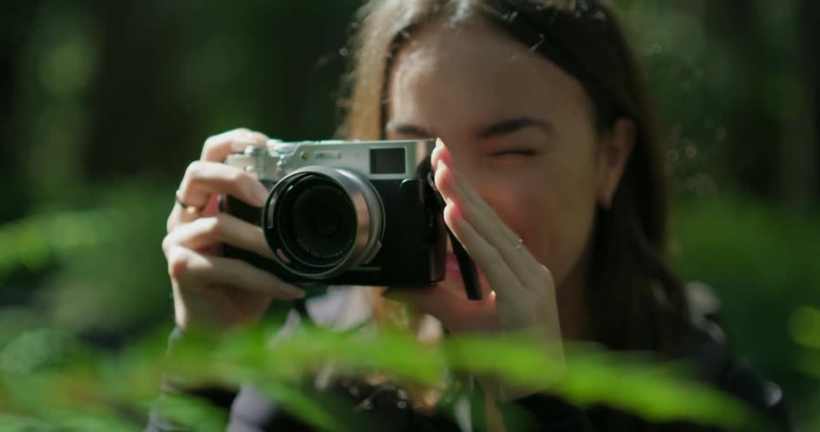 Woman with brown hair black shirt and backpack takes photo with camera smiling in the green forest