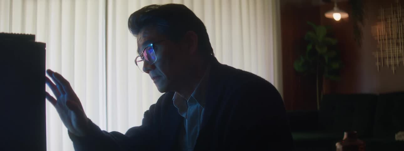A man with glasses sits looking concerned at a box and then raises his hand to touch it