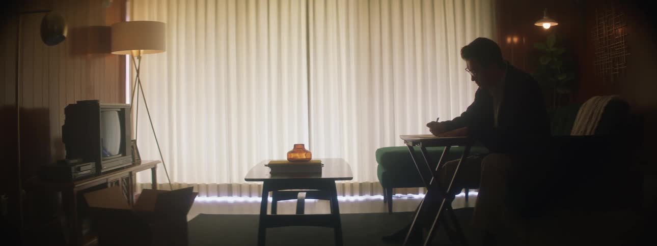 A man sitting on a chair using a table to write on with light coming through the window