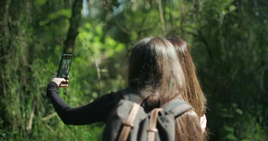 A woman standing in the woods wearing a backpack and holding her phone is standing next to another woman as she takes their picture together