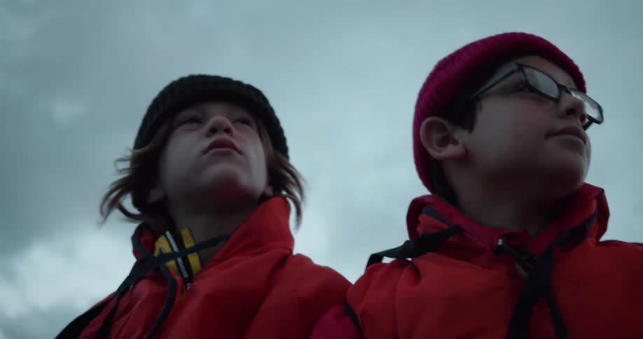 A boy with glasses looks around sitting next to another child who also looks around