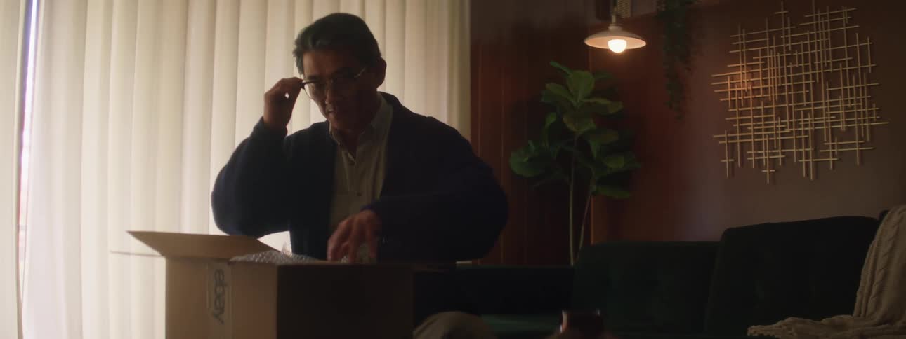 A man is sitting in a home while opening a large cardboard box in the daytime