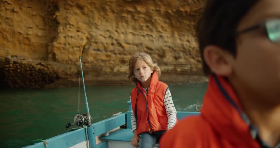 A little boy with dark hair and glasses in the front of a little blonde boy sitting behind him and a man standing on the boat