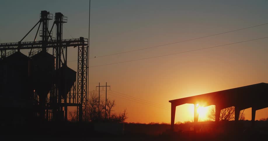 Factory tanks and buildings are silhouetted against sun sitting on horizon in red sky