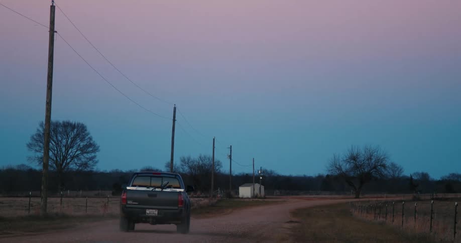 A pickup truck driving down a dirt road against a pink and blue sunset