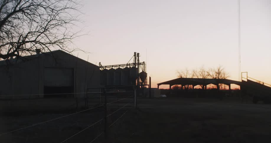 A still shot of a barn is shown on a flat grassy desolate area with a wire fence and a large sky background