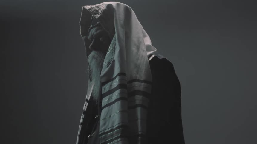 A man with a white beard wearing a head scarf and robe holds a large rock