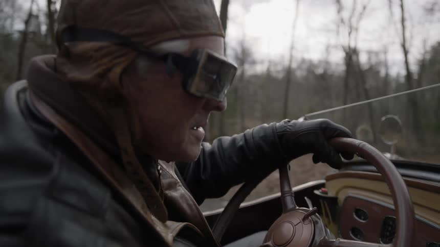 A man in cap and goggles drives an old convertible car