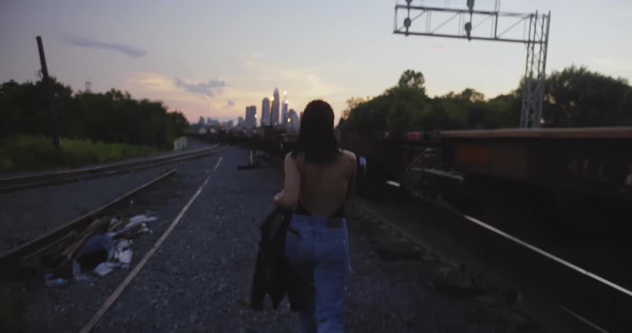 A woman removes her jacket while walking through the gravel beside a railroad as a train passes by