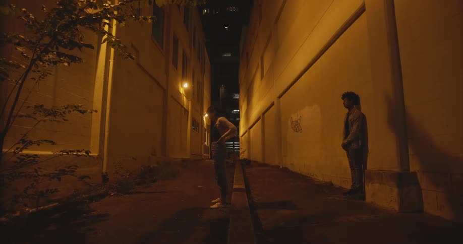 Girl sitting on vertical curb stands up and walks up to boy leaning against the wall in the alley at night