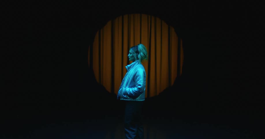 Woman tinted blue by lighting stands in spotlight near stage curtain