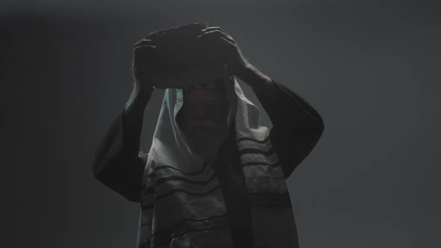 A man with a white beard wearing a headscarf and robe lifts a large rock above his head and strikes something below him