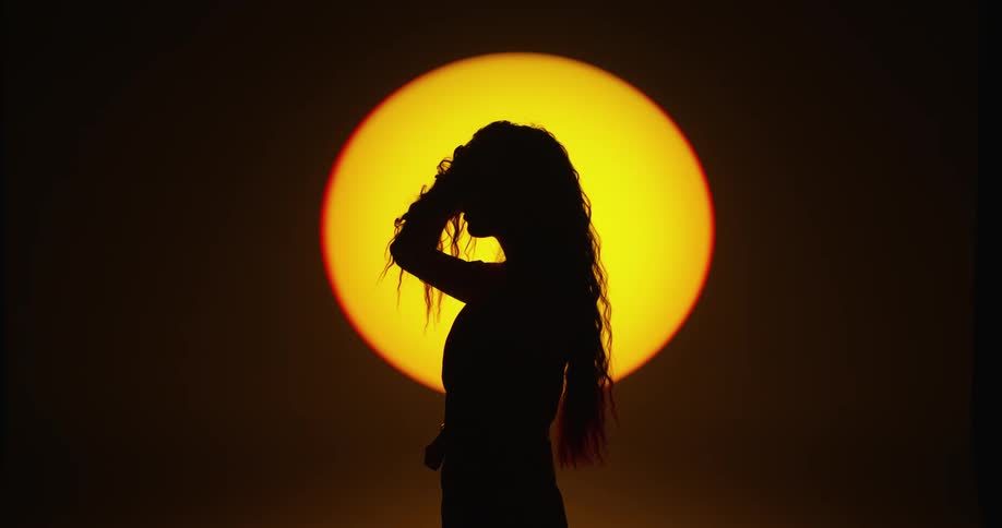 Side view silhouette of a woman moving her hand through her long hair in front of a round yellow light