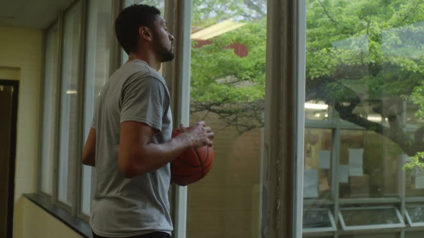 Black man holding a basketball looking out window contemplating