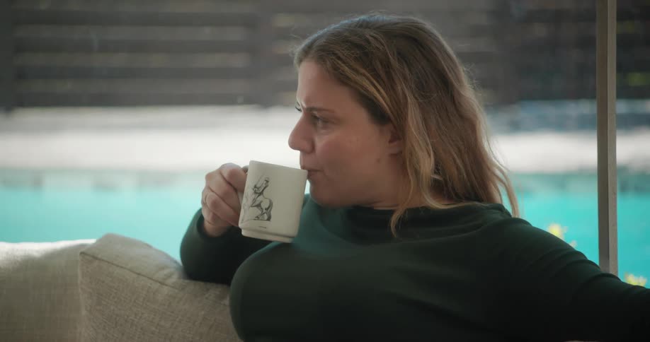 Woman drinks from a mug while sitting on couch near sliding glass doors showing a pool
