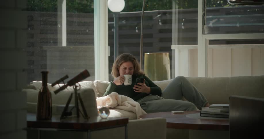 Woman sitting on the couch during the day using her smartphone and drinking a cup of coffee