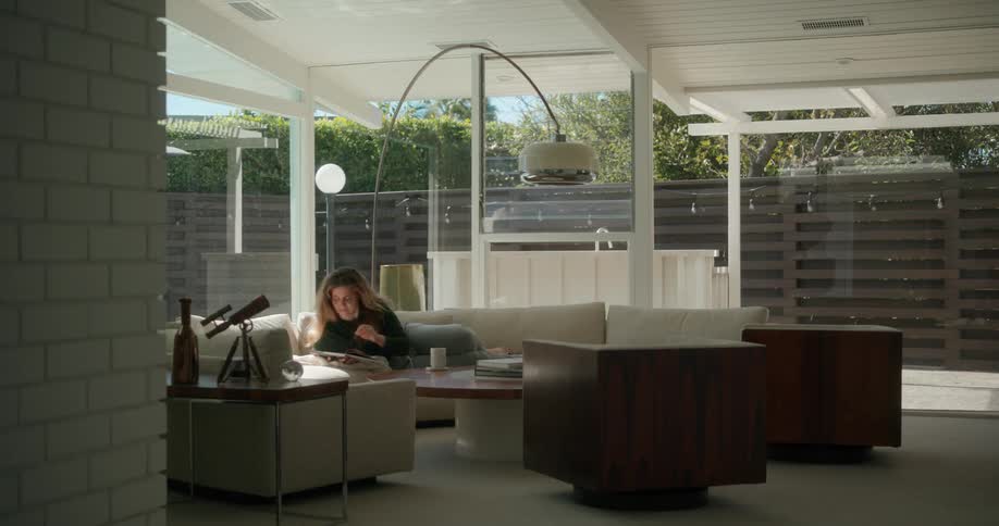 A woman is at home laying on the couch in a lavish living room sipping from a mug and flipping through a magazine