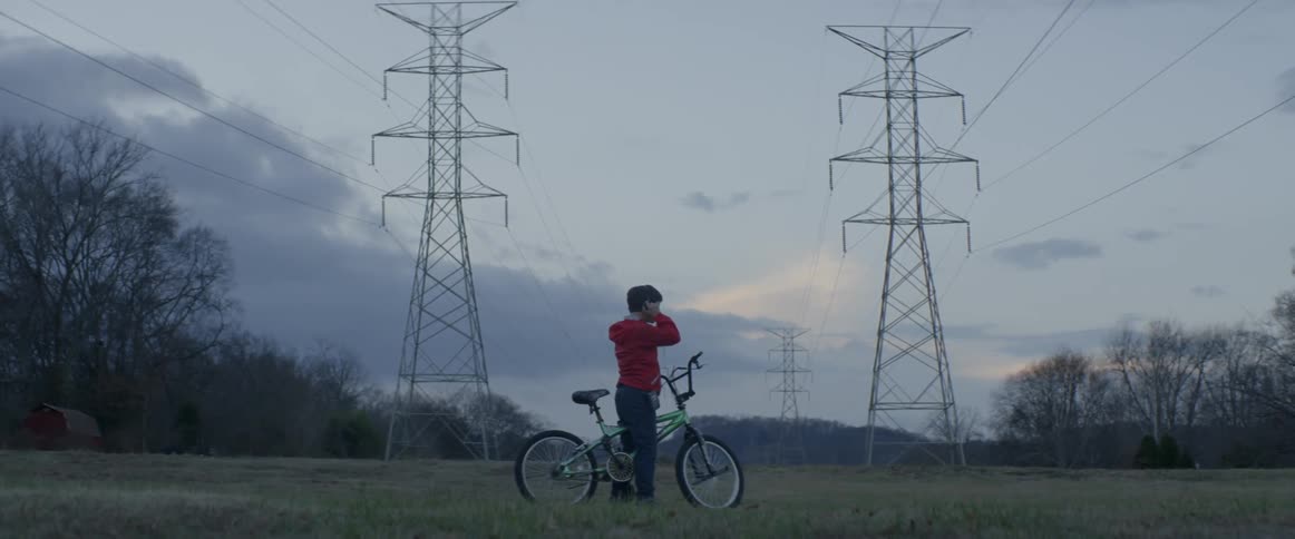 A young boy rides his bicycle near power towers and stops to put his headphones on