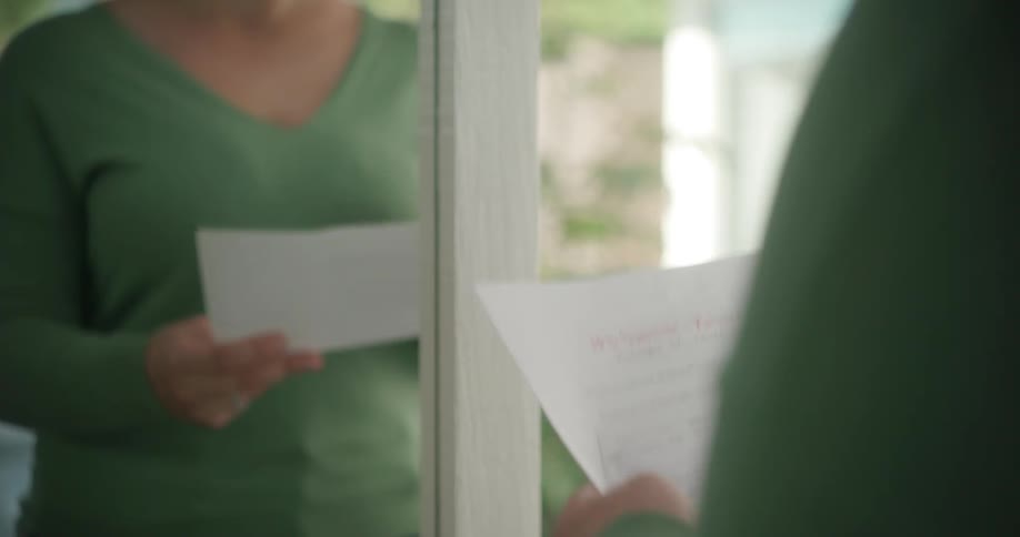 A woman stands at home and reads a paper as she crosses items off