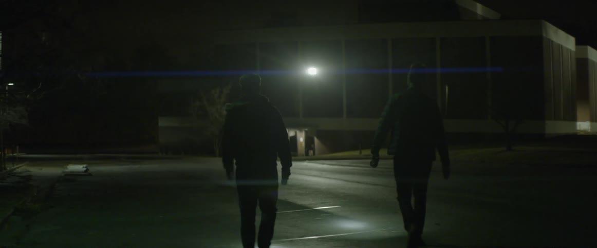 Two men holding flashlights walk toward a building
