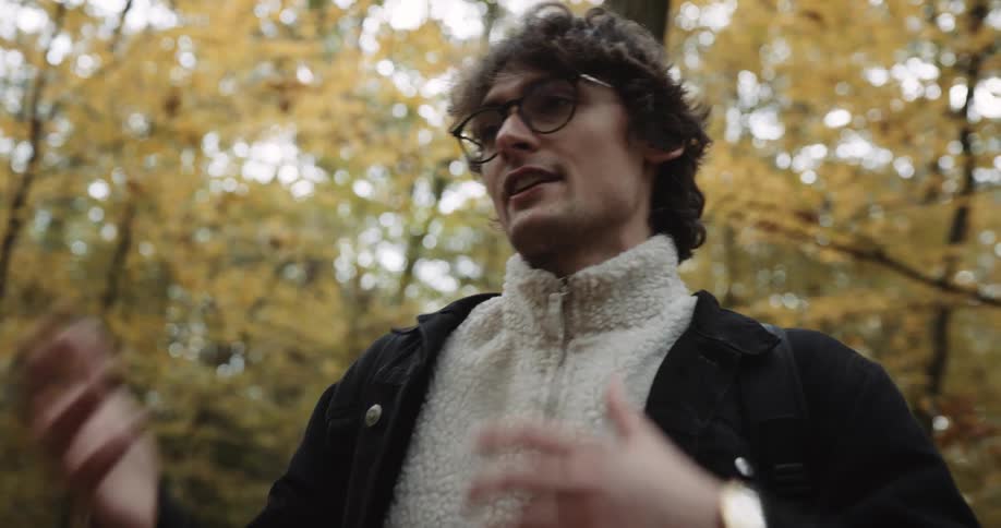 A man is standing in the woods moving his hands and arms as he speaks