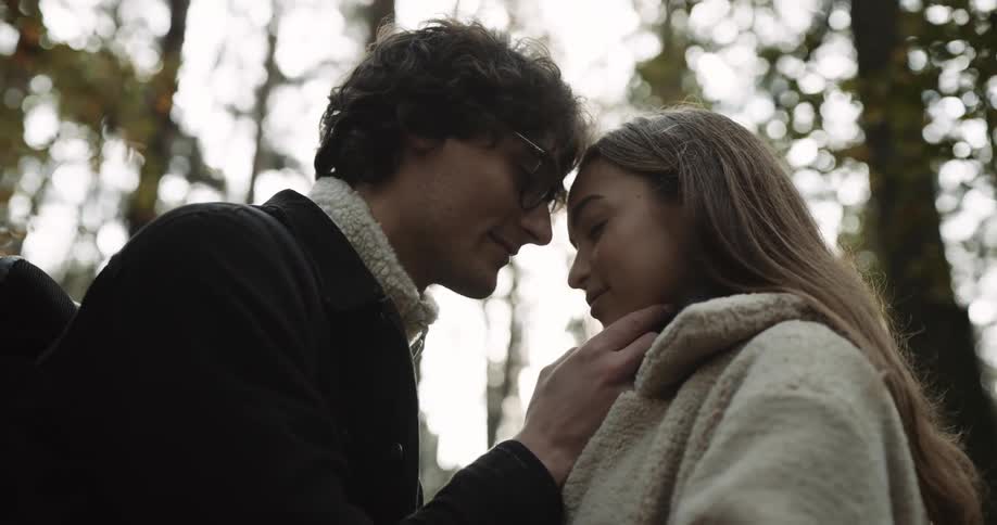 Man with glasses black coat and backpack touches woman wearing brown hair and beige coat in the forest
