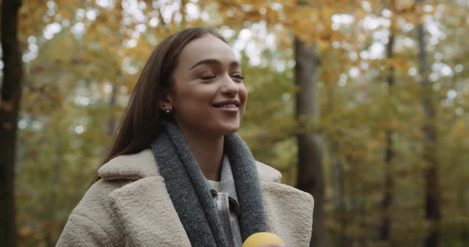 Woman with brown hair beige coat and gray scarf stands in the autumn forest listening and talking