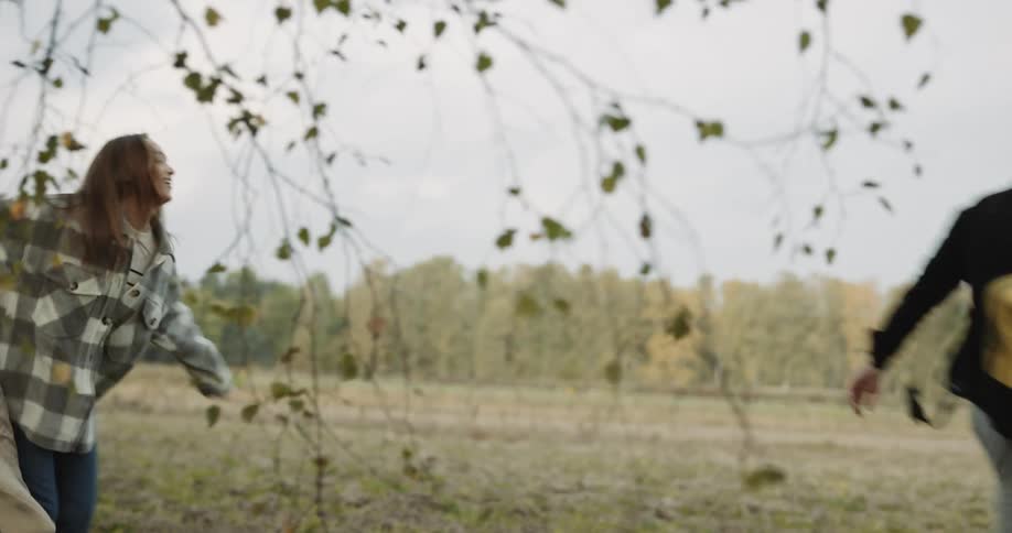 A woman chasing a man through trees playfully then jumping into his arms and both falling to the ground