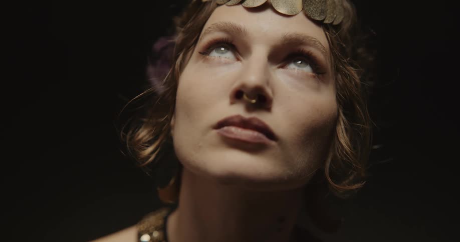 An up close of a womans face wearing a nose ring and head jewelry looking upwards and then at the camera