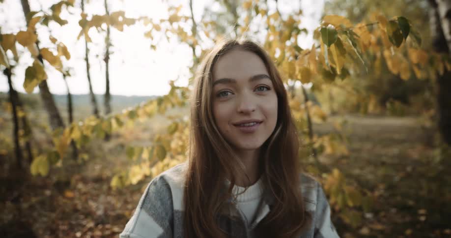 Woman with brown hair white gray jacket jeans and a pair of brown boots sits on ground posing for the camera in the autumn forest at dusk