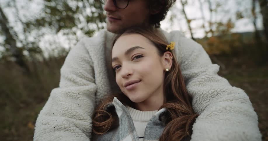 A woman grins and looks into the camera while sitting within the embrace of a man in a woodland during the day