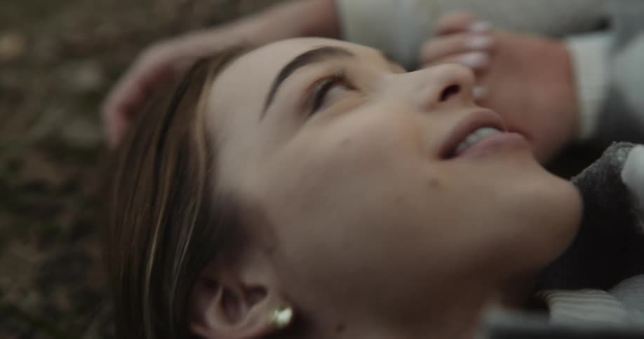 Woman with brown hair lies on ground next to man talking with each other in the forest
