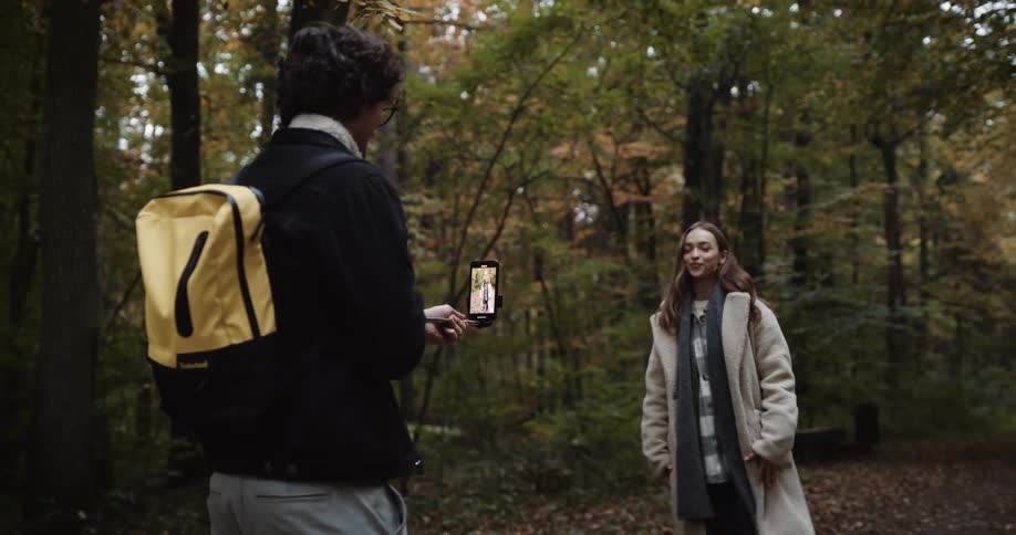 A teenage couple are hiking through the woods and he is holding his cell phone on a stick as he takes her picture
