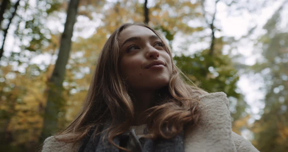 Woman with brown hair beige coat and gray scarf nods her head and looks down at camera in the autumn forest smiling