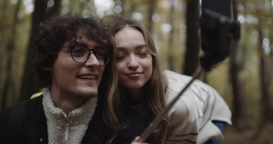 Man with glasses black coat and backpackand woman with brown hair and sweater take selfie with smartphone in autumn forest