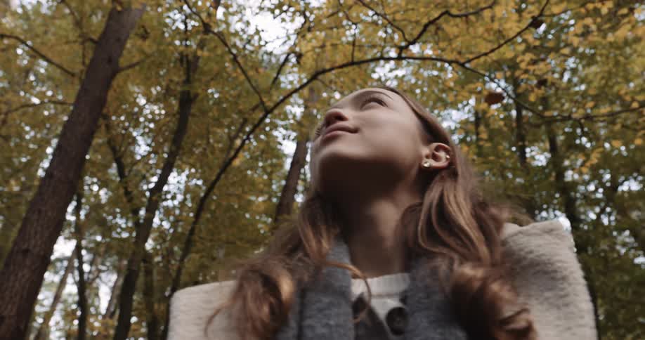 A teenage girl is standing in the woods looking around as autumn colored leaves fall from the trees