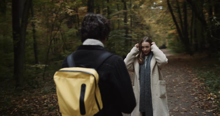 A teenage couple are hiking through the woods and he is holding a cell phone on a selfie stick as he takes her picture